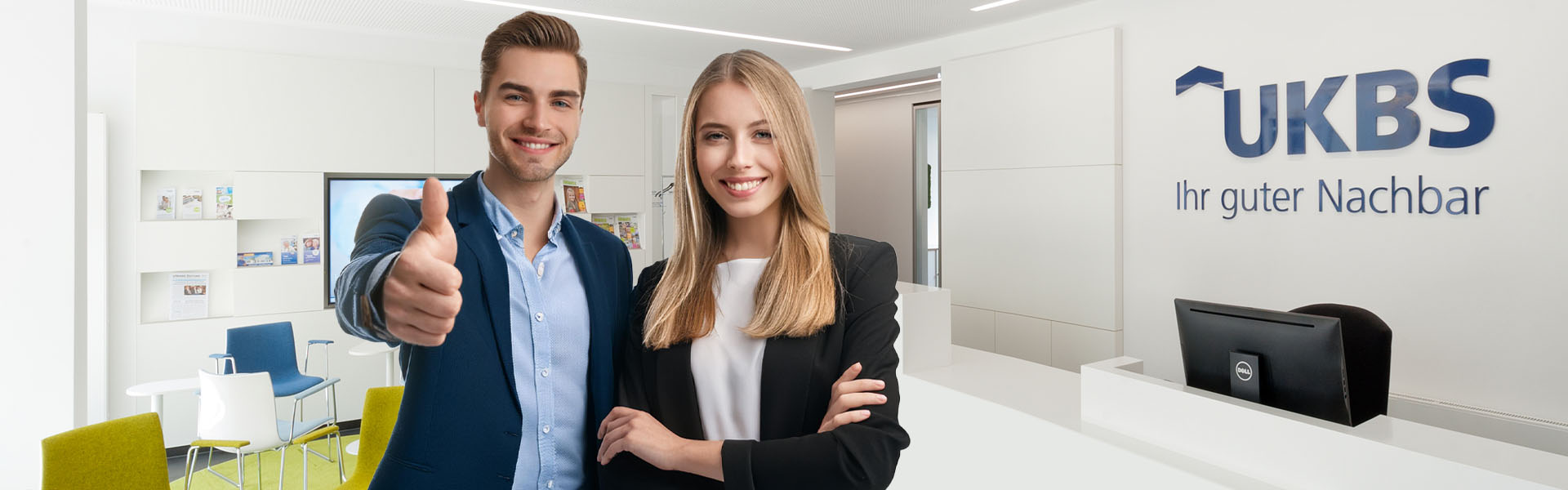 Ein junger Mann und eine junge Frau stehen im UKBS-Empfangbereich und lächeln den Betrachter freundlich an. Der junge Mann hebt den Daumen als Zeichen für eine Bewerbung bei der UKBS.